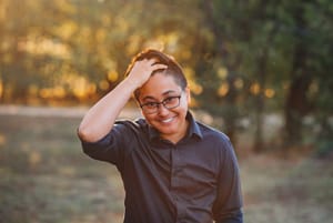 Nina smiling, wearing a dark blue shirt and black glasses, running hand through hair, backlit with blurred green background.